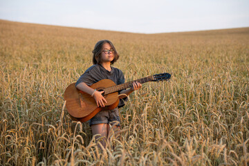 Naklejka na ściany i meble A girl in glasses with an old acoustic guitar stands with her eyes closed in a wheat field lit by the rays of the setting sun