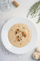 Top view of cream of mushroom soup made of pureed champignon with dairy cream decorated with fried slices and fresh thyme served in round white plate with glass of wine on wooden table. Vertical
