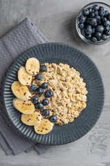 Top view of oatmeal is oat porridge made from oats decorated with fresh blueberries and sliced banana served in dark gray plate for healthy breakfast on concrete background with textile. Vertical