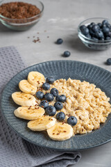 Oatmeal is oat porridge made from oats decorated with flax seeds, fresh blueberries and sliced banana served in dark gray plate for healthy breakfast on concrete background with textile. Vertical