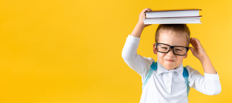 Banner Funny Preschool Child Boy in Glasses with Book on Head and Bag on Yellow Background Copy Space. Happy smiling kid go back to school, kindergarten. Success, motivation, winner, genius concept.