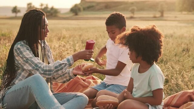 A good-looking afro-american mother and her children are eating hot-dogs while using ketchup sauce during a picnic outside at nature