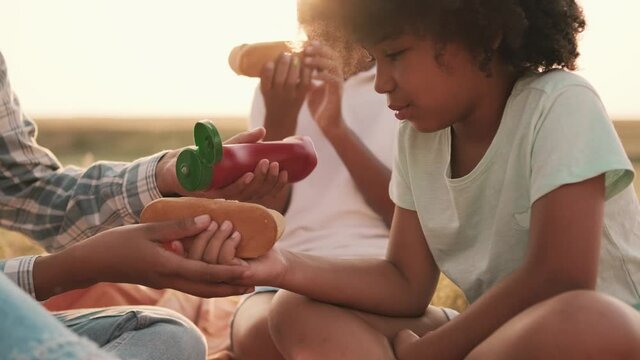 A close-up view of an afro-american mother and her children are eating hot-dogs while using ketchup sauce during a picnic outside at nature