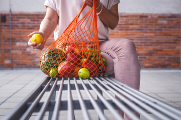 Woman with reusable mesh bag sitting on bench in city. Resting at bench after shopping fruits in supermarket. Zero waste and plastic free concept