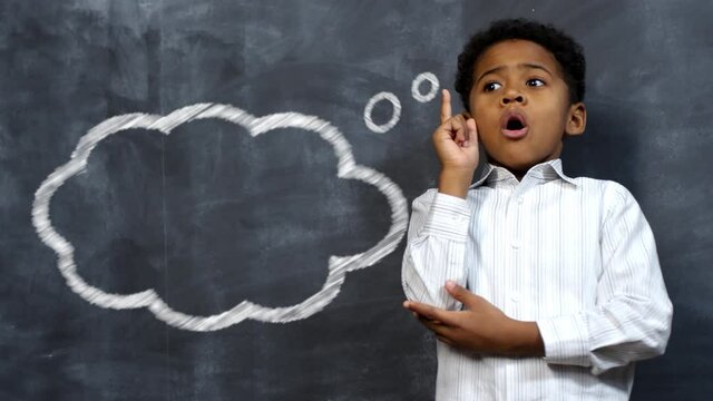 Little african american schoolboy opening mouth and pointing up with finger while standing against blackboard with computer animated chalk drawing of thought cloud