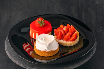 Three cakes on black plate. Fruit desserts with fresh strawberries. Confectionery. Delicate desserts