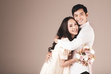 Portrait of young attractive Asian couple, man wearing white shirt, woman wearing beige dress hugging each other, woman holding bouquet of flower. Concept for pre wedding photography