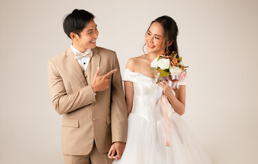 Young attractive Asian couple, man wearing beige suit, woman wearing white wedding gown standing...