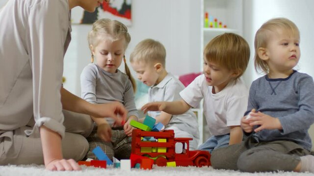 Cute little boys and girls putting color blocks into toy truck with help of female teacher while playing together in daycare center