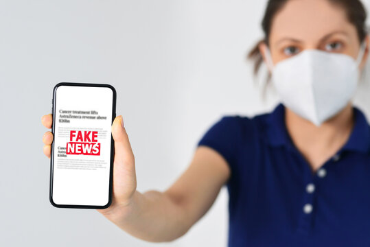 Woman Wearing Medical Mask And Holding A Smartphone With Fake News In Front. Propaganda, Misinformation. Horizontal Photo. Online Fake News On A Mobile Phone. 