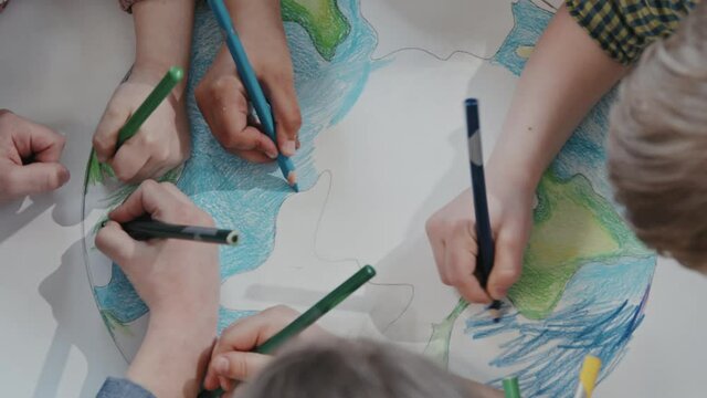 Handheld top view with close up of kids using pencils and coloring drawing of planet Earth