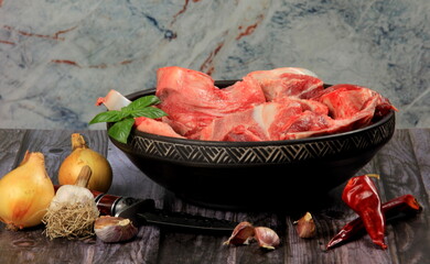 Raw beef bones in round clay bowl on wooden table decorated with vegetables and spices, vintage hunter knife. Beef selection for broth