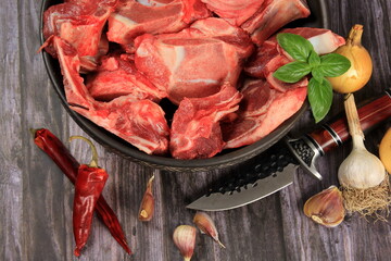 Raw beef bones in round clay bowl on wooden table decorated with vegetables and spices, vintage hunter knife. Beef selection for broth