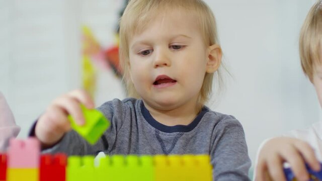 Rack Focused Shot Of Adorable 3 Year Old Boy Building Toy House With Colorful Plastic Bricks While Sitting At Table With Children And Playing With Construction Toy In Kindergarten