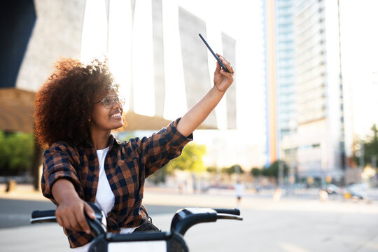 Young lady on electric bicycle on the street. Beautiful african girl taking selfie photo.