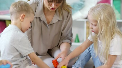 Tilt down of cute little boy and girl sitting with female teacher on the floor in playroom and building construction toy with colorful plastic bricks