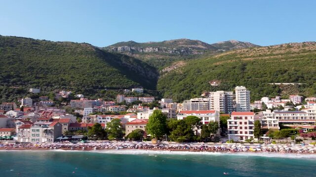 The drone flies parallel to the beach Petrovac, Montenegro. Blue waves crash on the shore, against the background of high mountains are the houses of the city