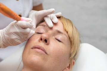 Close up portrait of young blonde woman in professional beauty salon getting permanent make up done in artificial light by cosmetologist in latex gloves during covid 19 pandemic