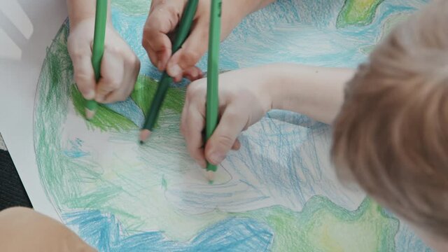 Handheld close up shot of hands of kids using pencils and coloring drawing of planet Earth