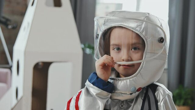 Handheld close up portrait of cute 6-year-old girl in spacesuit putting her visor down and looking at camera. Cardboard rocket ship is in background
