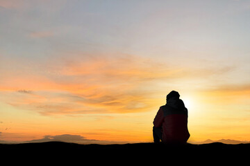 Silhouette of young man sitting and watching beautiful view overcast sky, sunset alone on top of the mountain. He enjoyed traveling and was successful when he reached the summit.
