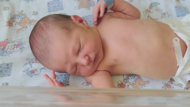 A newborn baby sleeps restlessly and shudders behind a transparent glass in a maternity hospital bed, a close-up portrait