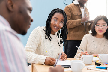Woman in glasses with dark complexion and pigtails sits in office at work in conference meeting with colleagues, discussing business project, filling out documents, drinking coffee, corporate work