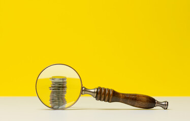stack of coins and a wooden magnifier on a white table, yellow background. The concept of analysis...