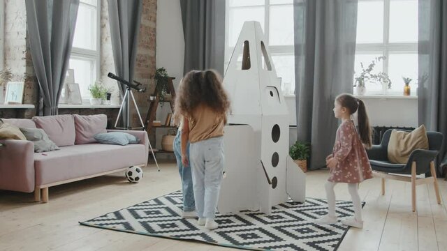 Handheld shot of group of cute little kids playing with cardboard rocket ship model at home