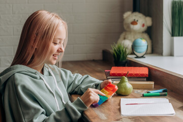 Fototapeta na wymiar Positive blonde teenage girl playing with colorful pop-it fidget toy while studying at home.Trendy anxiety and stress relief fidgeting game.School concept.