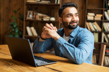 Happy and optimistic Indian man in casual wear using laptop in modern office, successful inspired...