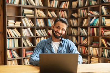 Cheerful Indian male freelancer looks at the laptop screen, eastern student websurfing sitting at...