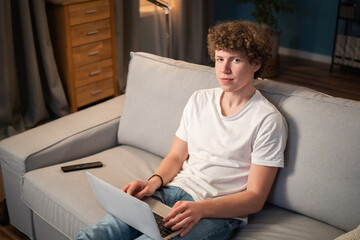 A young boy with curly hair sits on the living room couch with his laptop in his lap and proudly stares into the camera.