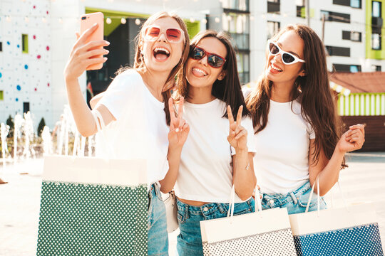 Three young beautiful smiling hipster female in trendy summer same clothes. Sexy carefree women posing in the street.Positive models having fun in sunglasses.Holding shopping bags.Taking selfie photos
