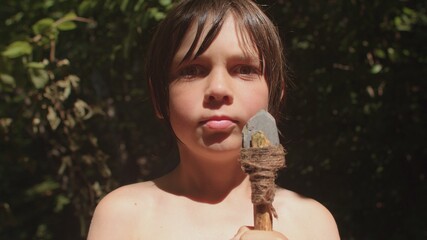Portrait of a boy with a handmade spear in the forest. Long haired preteen playing survival game, explore, master crafting. The spot of the sun illuminates the face. The stone tip is tied with a rope