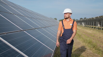 Male engineer in uniform walking and looking at solar power plant. Man in hard helmet examining object. Concept of solar station development and green energy. Worker on solar power station outdoor