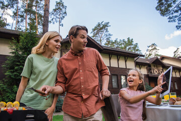 Smiling kid pointing at digital tablet near parents and grill outdoors