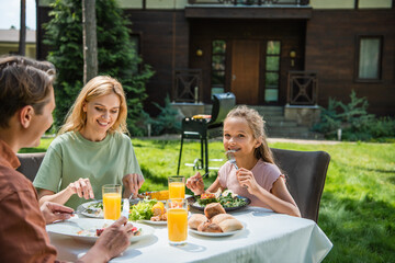 Smiling kid looking at camera while eating salad near parents during picnic