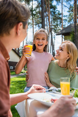 Smiling girl holding orange juice near mother and father during picnic and vacation