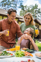 Happy parents with orange juice looking at daughter near tasty food during picnic