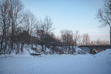 ice and snow covered river in Latvia in cold winter evening. Bridge in distance. Old clay brick house on left shore, there are trees and footbridge too