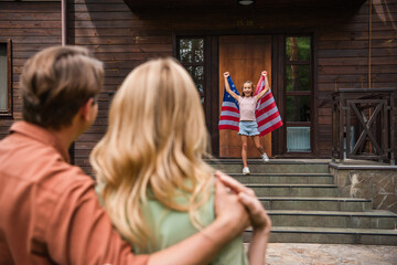 Fototapeta na wymiar Excited girl holding american flag near blurred parents embracing outdoors