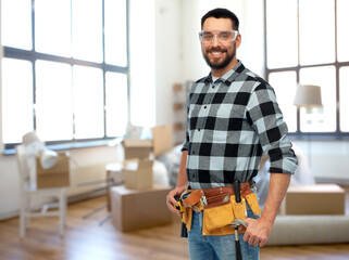 repair, construction and building concept - happy smiling male worker or builder in goggles with tool belt over home room background