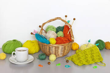 Needlework and knitting as a hobby in autumn. Wicker basket with balls and knitting needles and skeins of woolen yarn on table. Still life with a cup of green tea and knitting accessories. DIY concept