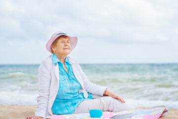 A very old wrinkled pensive woman sits on the seashore with a cup of tea and a tablet.