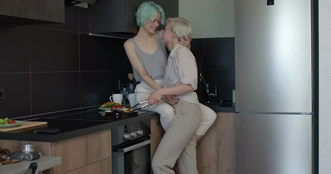 two cute lesbian female friends making breakfast in kitchen, kissing and hugging. people lifestyle. Side view on caucasian two female in casual domestic clothes spending weekends morning together