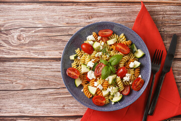 Italian pasta fusilli tricolore with cherry tomatoes, zucchini and feta cheese in a blue shallow bowl on a red napkin with black silverware on a wooden surface