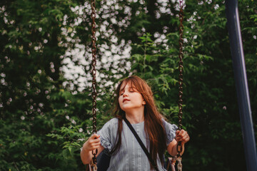 Preteen girl wear blue eco cotton dress enjoy swing in summer park with lush green trees. Sincere...