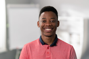 Portrait of young boy with dark eyes, hair, black skin, beautiful wide white smile, blurred background, man is wearing salmon collar shirt, student in class