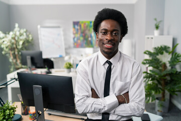Fototapeta premium Smiling friendly dark skinned afro men sits on desk wearing white shirt and tie, in background scenery of company, office boss, president, manager, businessman, arms crossed on stomach